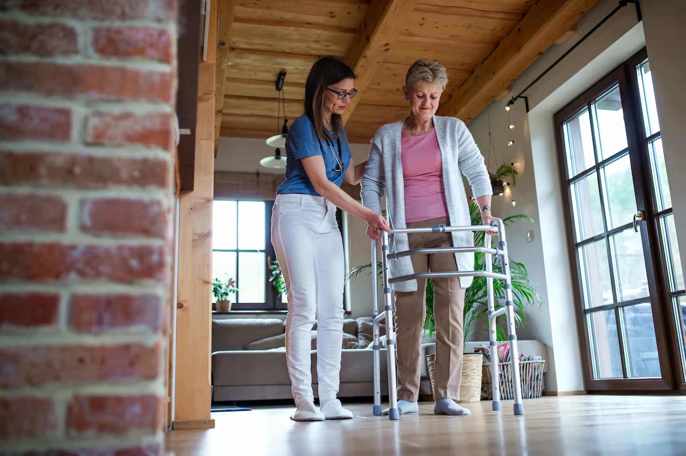 An older woman walking with a frame at home, steadied by her carer